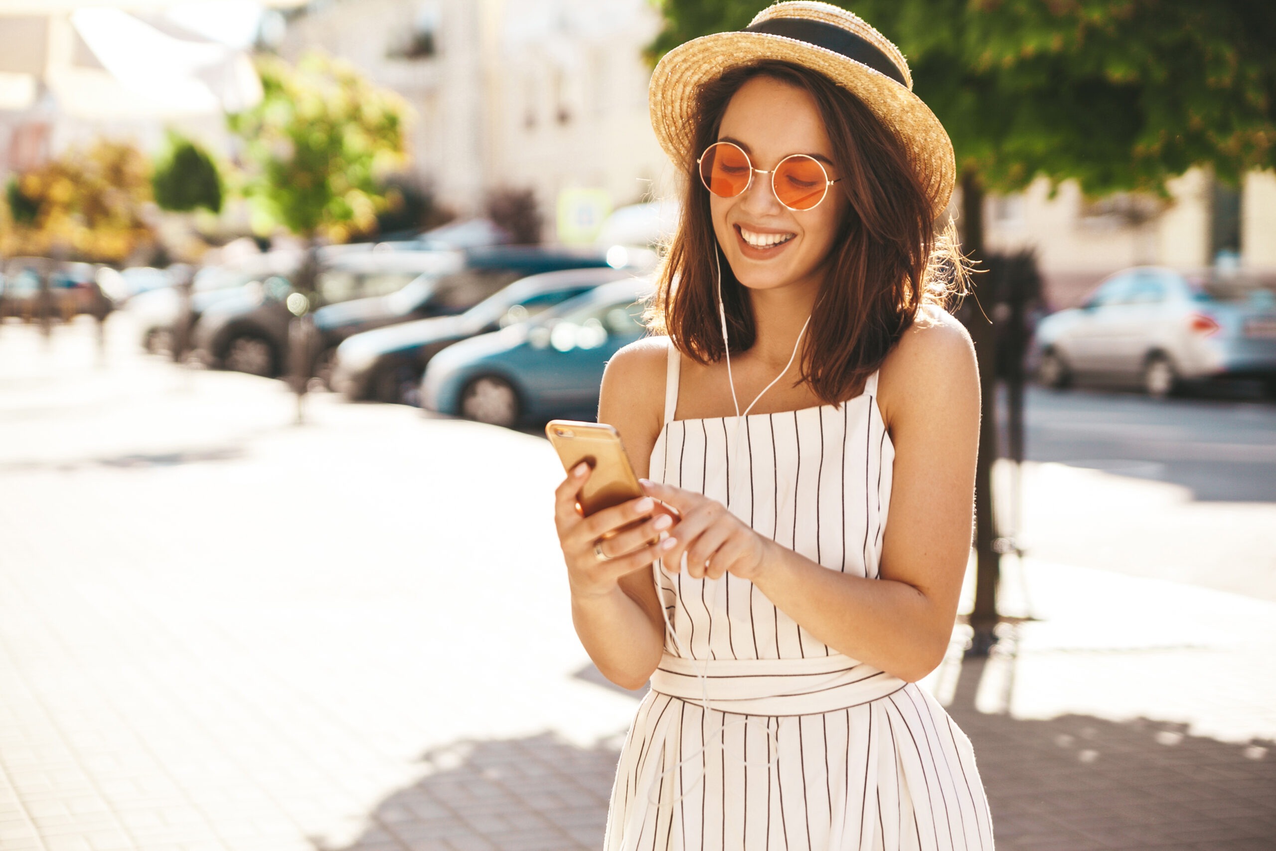 Portrait of happy smiling teen girl in summer hipster clothes listening music on her headphones from smartphone in the street on a sunny day